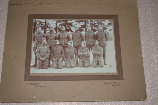 Sepia photograph of 11 men six standing and five kneeling dressed in military uniform wearing forage cap with row of pine trees in background.  Photograph is mounted on brown cardboard with handwriting across top border.