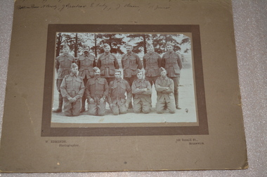 Sepia photograph of 11 men six standing and five kneeling dressed in military uniform wearing forage cap with row of pine trees in background.  Photograph is mounted on brown cardboard with handwriting across top border.