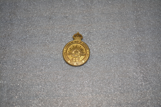 Gold coloured metal badge round in shape with rising sun in centre and crown on top