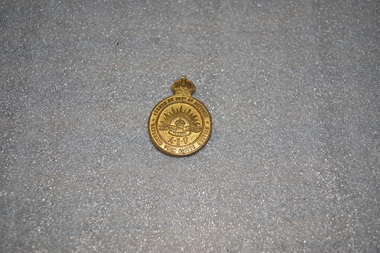 Gold coloured metal badge round in shape with rising sun in centre and crown on top
