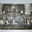 Black and white unframed photograph of large group of men and women in formal dress front of stage with flags in background