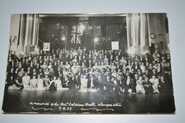 Black and white unframed photograph of large group of men and women in formal dress front of stage with flags in background