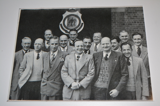 Black and white unframed group photograph of 14 men standing under RSL Logo shield.