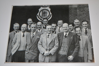 Black and white unframed group photograph of 14 men standing under RSL Logo shield.