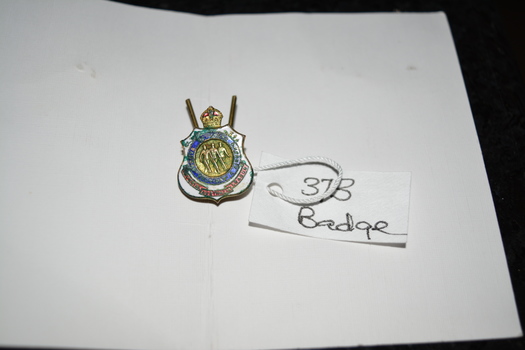 Gold badge with crown on top, blue circle in the middle with image in gold centre, red banner at bottom surrounded in white enamel.