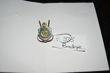 Gold badge with crown on top, blue circle in the middle with image in gold centre, red banner at bottom surrounded in white enamel.