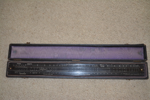 The long narrow case is fabric covered wood with felt fabric lining containing a long black laminate ruler with numerous white markings and numbers.  Top of the ruler is in three sections with the centre section movable by small gold metal knobs at either end.
 