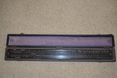 The long narrow case is fabric covered wood with felt fabric lining containing a long black laminate ruler with numerous white markings and numbers.  Top of the ruler is in three sections with the centre section movable by small gold metal knobs at either end.
 
