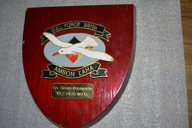 White seagull in flight over blue background beneath and above two scrolls mounted on shield shaped timber plaque