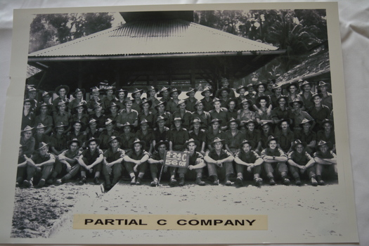 Large group of soldiers half sitting and half standing in fron of building with corrugated roof.