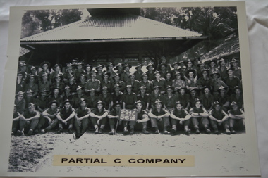 Large group of soldiers half sitting and half standing in fron of building with corrugated roof.