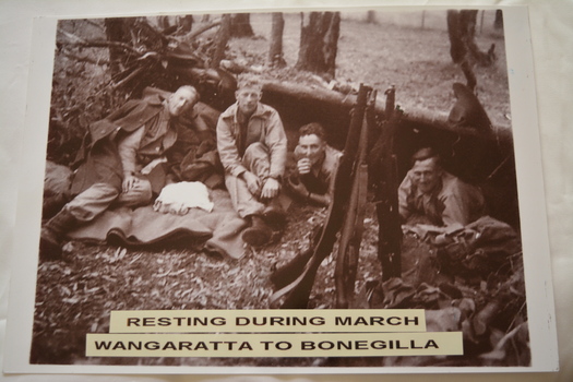 Four soldiers resting in various positions on the ground and four rifles  pitched upright to form a tent in the foreground