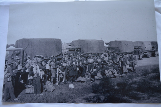 Large group of soldiers with packs/kit standing in front of row of trucks with canvas covers