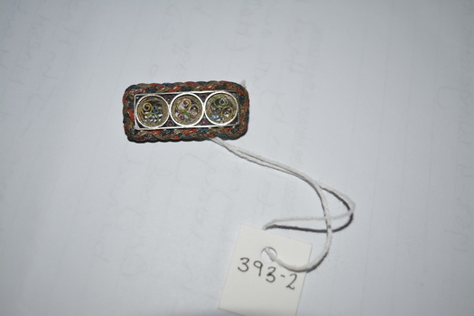 Rectangle metal brooch housing three filigree metal circles with red and blue plaited metal surround.