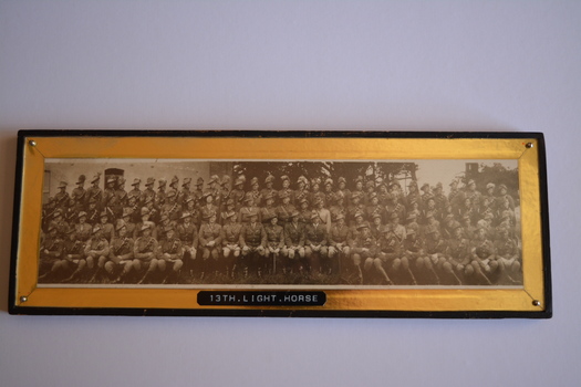 Wide and narrow rectangle shaped black painted frame with gold colour mount and four silver metal screws in each corner. The frame contains a black and white photograph depicting a large group of men dressed in military uniform with bandoliers and slouch hat, some with emu plumes.