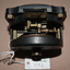 Black painted cylindrical instrument with glass face .  There is a side cutout containing  metal parts .