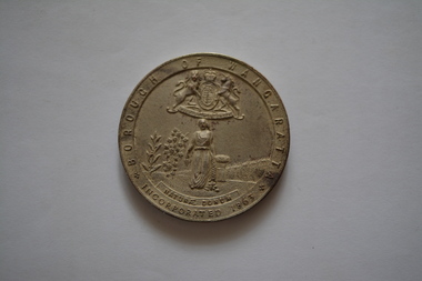 Circular silver metal medal- obverse side has coat of arms above image of woman in a field - rear side embossed with laurel wreath.