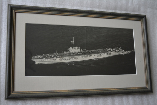 Framed black and white image of a ship with aircraft on deck at ship’s bow and stern.