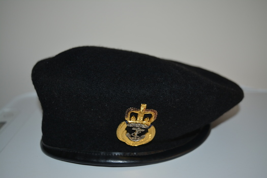 Black wool beret with black vinyl band.  At front two eyelets affixed with gold coloured metal badge consisting of an anchor below a crown.