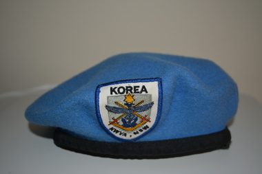 Blue wool beret with black braid band.  At front affixed with multi coloured fabric badge consisting of an anchor, boomerang, wings and crossed swords below a yellow star. 