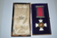 Box contains a crimson ribbon with dark blue edges with a suspension bar and pin at the top and a lower suspension bar attached by a loop to a cross shaped medal made from white enamel. 