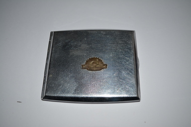 Square silver metal case with left side hinge. A small rising sun badge is affixed in centre on top cover.