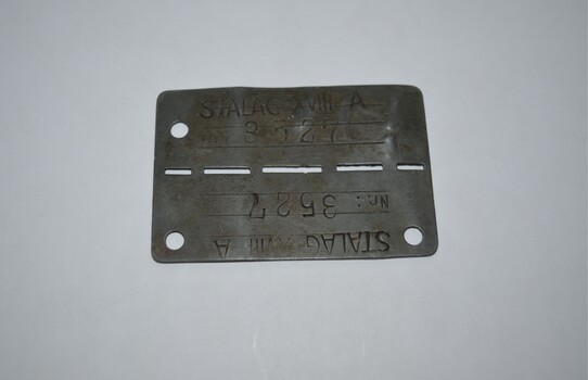 Dark grey rectangle shaped piece of metal with small hole in three of the four corners and narrow cut out slots across centre line stamped either side with letters and numbers.