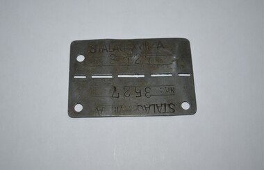 Dark grey rectangle shaped piece of metal with small hole in three of the four corners and narrow cut out slots across centre line stamped either side with letters and numbers.