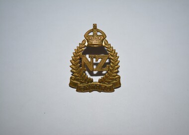 Gold colour metal badge takes the form of a laurel wreath surmounted by a crown. In the centre are the voided letters 'NZ'.