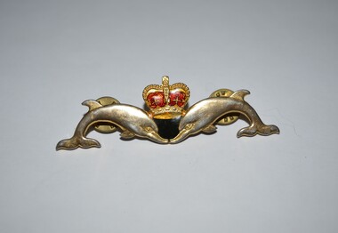 Gold plated metal badge consisting of a gold and red enamel crown between two dolphins.