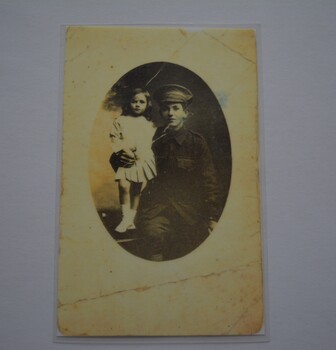 Laminated rectangle in shape containing oval black and white photograph of soldier sitting with his arm around little girl in white dress.
