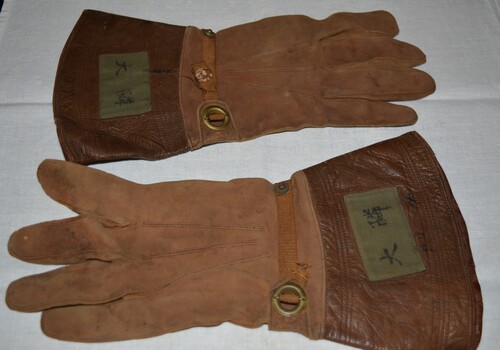Brown suede gloves with brown ribbon strap threaded through an eyelet and secured with a press stud.  