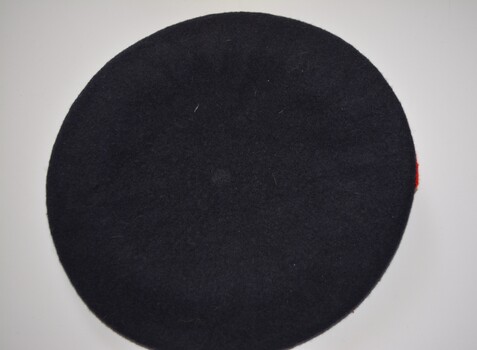 Dark blue felt beret with black leather trim with enclosed ribbon with white metal badge of coat of arms attached to red felt square handsewn at front.