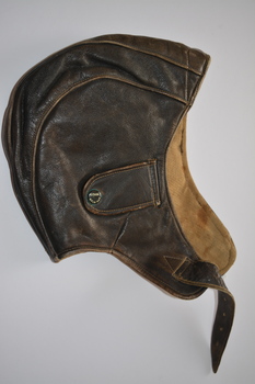 Brown leather flying helmet with buckle chinstrap and small buckled loop on the rear to retain goggles.  External earpiece openings are secured by a pair of snap fasteners with brown plastic caps.