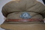  Brown peaked cloth cap with metal badge at front above narrow brown leather strap attached by brass buttons either side of peak.
