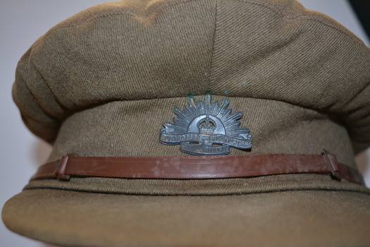  Brown peaked cloth cap with metal badge at front above narrow brown leather strap attached by brass buttons either side of peak.