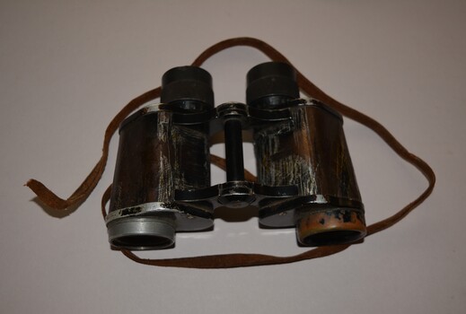 Black painted metal binoculars originally covered in black leather with little remaining.  The brown leather strap is well worn and missing stud fixing on right side.