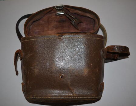 Brown leather look binocular case lined in brown colour cloth.  Long narrow leather strap fixed along sides by leather secured in place by metal studs.  Fitted lid is attached by leather strap fixed in place by four metal studs at rear.  At front is a spring closing mechanism which is broken.