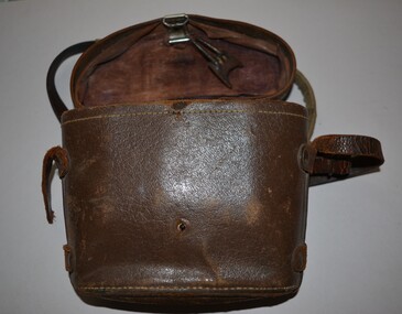 Brown leather look binocular case lined in brown colour cloth.  Long narrow leather strap fixed along sides by leather secured in place by metal studs.  Fitted lid is attached by leather strap fixed in place by four metal studs at rear.  At front is a spring closing mechanism which is broken.