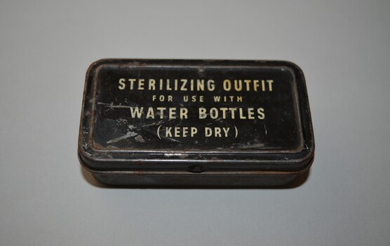 Rectangular tin with hinged lid containing two clear glass vials of water sterilization tablets. The tin is plain metal apart from the black lid with white lettering.  The two vials are stoppered  with cork. 