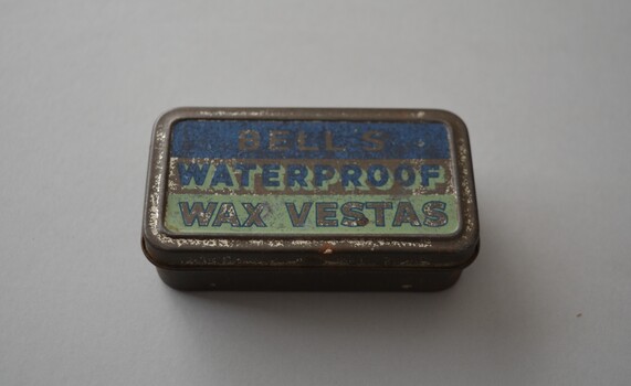 Rectangular tin with hinged lid containing small timber matches with wax heads. The tin is plain metal apart from two tone blue lid with lettering.   Underneath is a serrated patch used to strike the match.