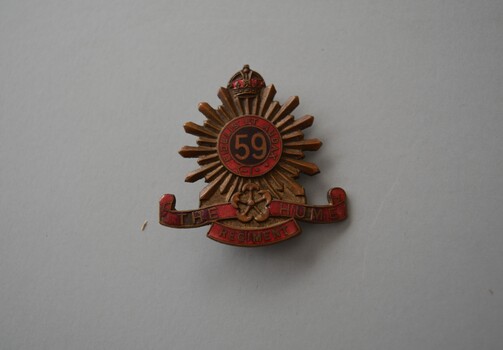 Brass badge features a central red enamel circle containing the raised numeral 59 circled by a motto with radiating design with crown on top and scroll underneath