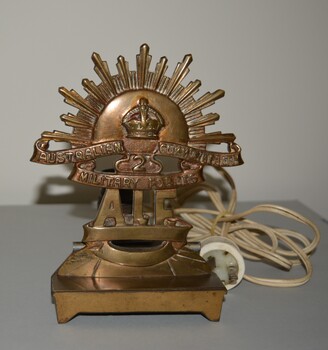 Brass Rising Sun Badge mounted on brass stand with light fitting attachment and white plastic electrical cord.