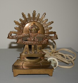 Brass Rising Sun Badge mounted on brass stand with light fitting attachment and white plastic electrical cord.