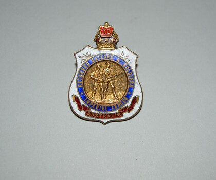 Shield shaped badge with crown on top. White enamel with blue circle in centre above red colour scroll. Image of two servicemen with rifles. 