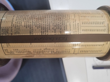 A cylindrical slide rule composed of a lacquered cylinder and a mahogany handle at one end. A brass pointer with an engraved index marker at its tip is attached to the handle and a second brass pointer is attached to the top cap. 