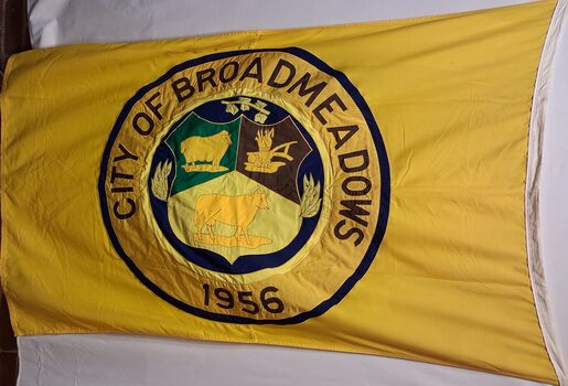 Yellow municipal flag featuring the City of Broadmeadows civic emblem within a blue-bordered circular seal. The emblem incorporates agricultural imagery and is surrounded by the inscription “CITY OF BROADMEADOWS” and the date “1956”. The flag displays creasing from folded storage and is photographed against a light background.
