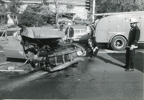 A wrecked car centre left with three firemen looking at the car, a fire engine in the background on the right.