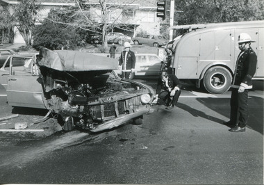 A wrecked car centre left with three firemen looking at the car, a fire engine in the background on the right.