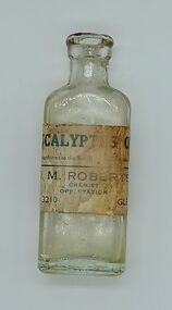 Glass bottle with a blue and white label. The label shows the contents were eucalyptus oil and was sold at D. M. Roberts, Chemist, sited opposite the station.